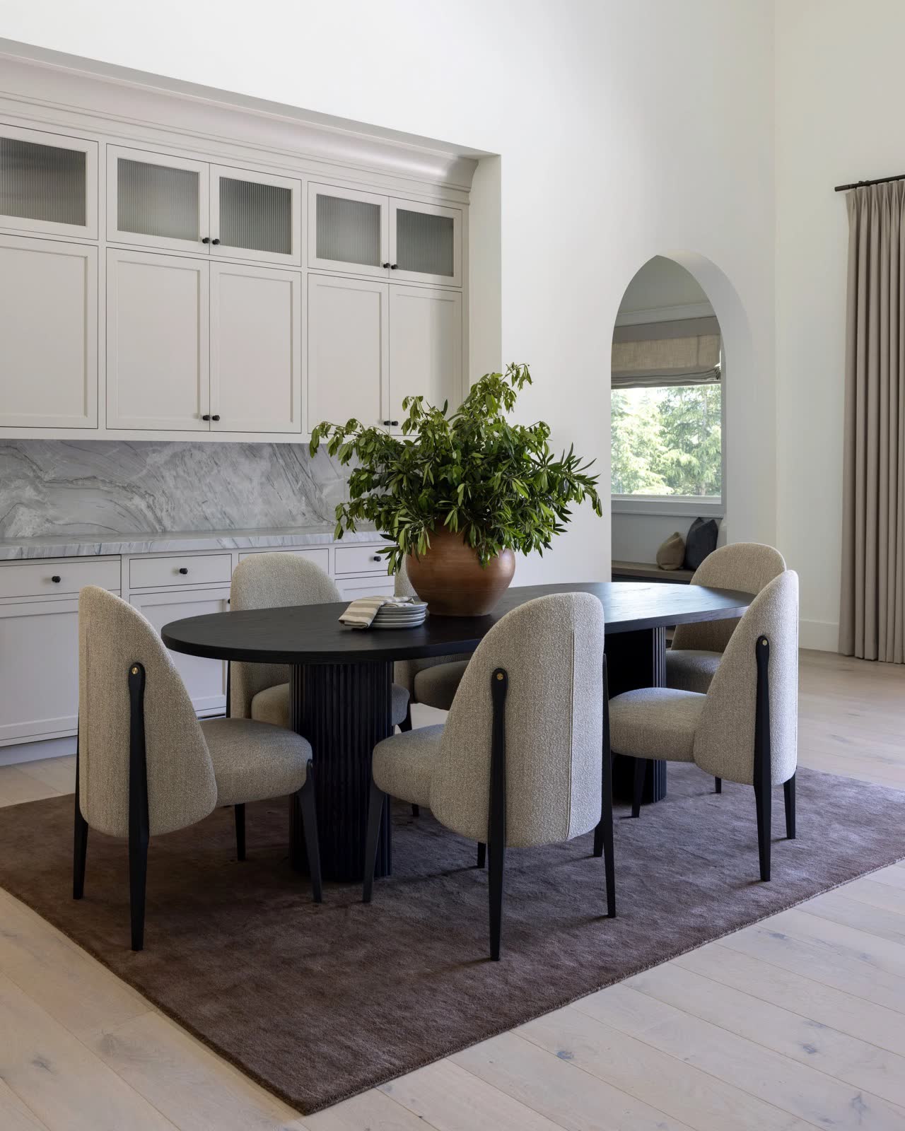 A dining room with a black oval pedestal table, cream chairs, and a marble buffet.