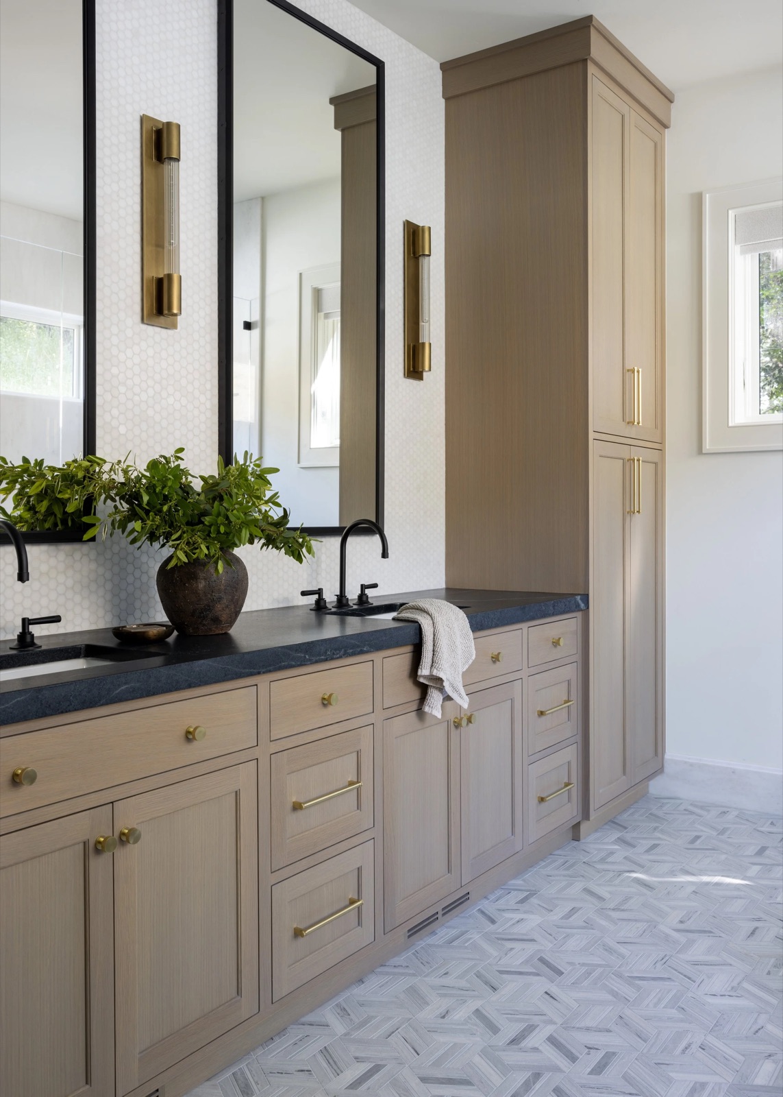 A bathroom vanity in warm oak with brass sconces, brass pulls, and a dark stone counter.
