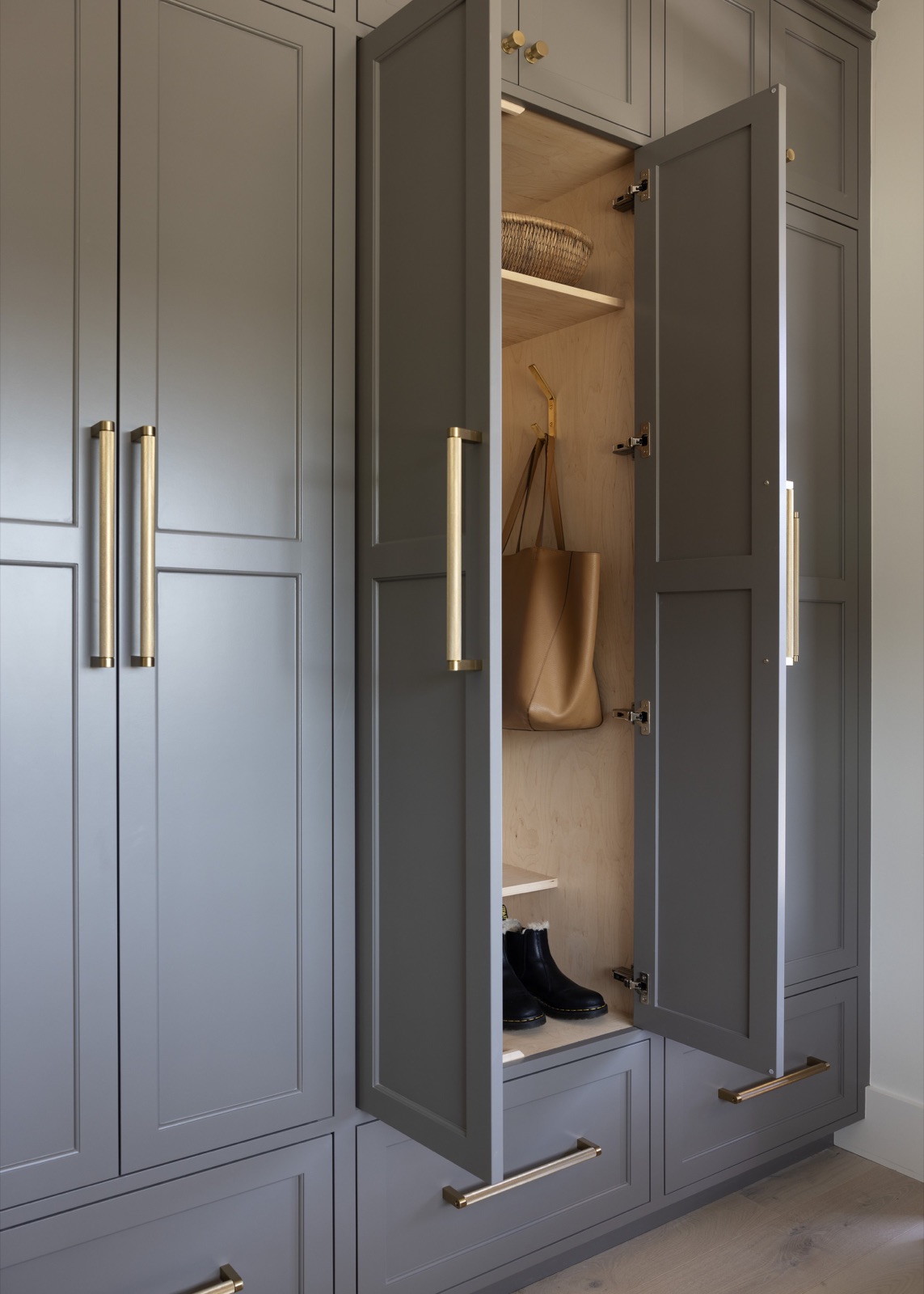 Floor-to-ceiling mudroom cabinets in warm gray, opened to a maple interior with brass hardware.