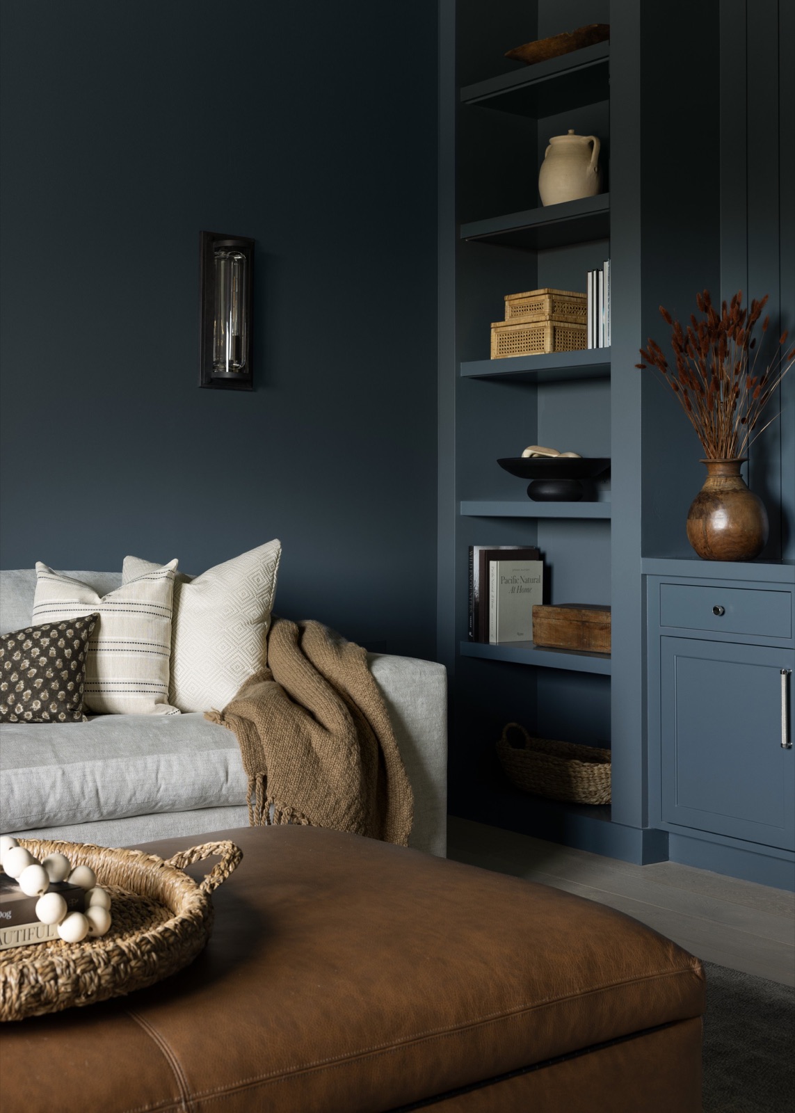 The den, deep blue walls and built-in shelving around a cream sofa and leather ottoman.