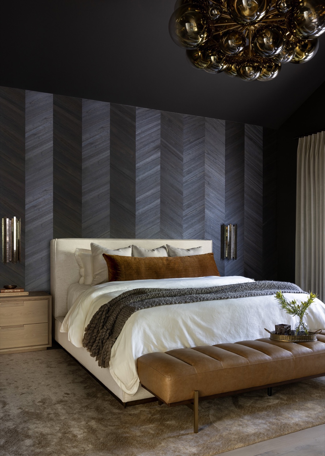 The primary bedroom, a dark chevron wood feature wall, brass bubble chandelier, and leather bench.