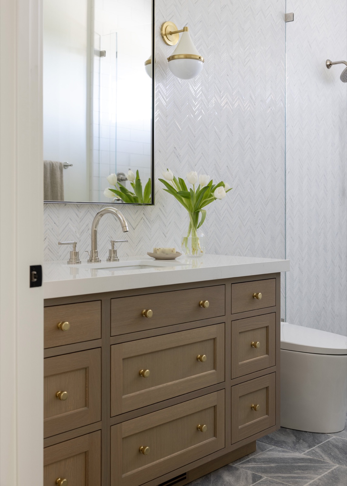 A bath vanity in rift oak with brass knobs, beneath a herringbone marble tile wall.
