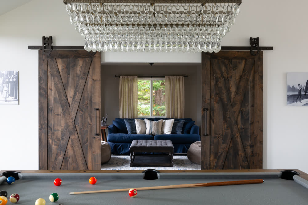 The game room, a pool table beneath a glass chandelier, barn doors opening to the lounge.