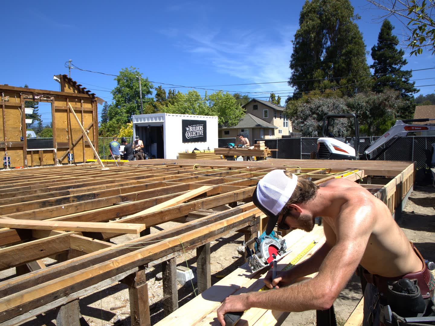 A Builders Collective carpenter cutting lumber on site, with new floor framing behind him.